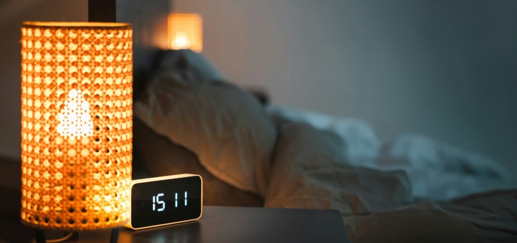 A digital clock sitting on a nightstand next to a turned on lamp